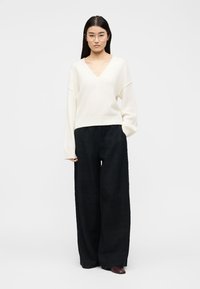 Γυναίκα με διάφανα γυαλιά, λευκό πουλόβερ με λαιμόκοψη σε σχήμα V, φαρδιά μαύρη παντελόνα και καφέ παπούτσια, στέκεται με το ένα χέρι στην τσέπη.