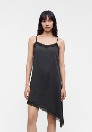 Femme portant une nuisette noire en satin avec bordure en dentelle, fines bretelles et ourlet asymétrique sur un fond blanc uni.