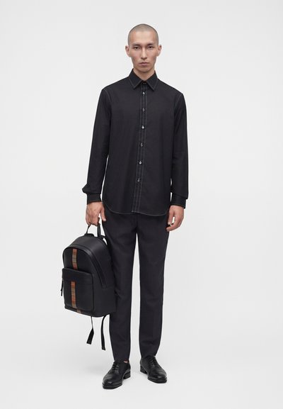 Homme portant une chemise noire boutonnée et un pantalon, tenant un sac à dos noir avec un détail rayé, debout devant un fond blanc.