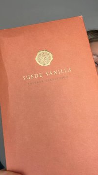 Hardcover bok i ferskenfarge med tittelen "Suede Vanilla" med tekst i gullpreget skrift og en sirkulær emblem øverst. Teksturert finish på omslaget.