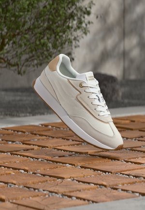 Άσπρο και μπεζ χαμηλό sneaker με κορδόνια, που αιωρείται πάνω από ξύλινο πλακάκι δαπέδου με χαλίκι ανάμεσα στα πλακάκια, πράσινο φύλλωμα στο παρασκήνιο.