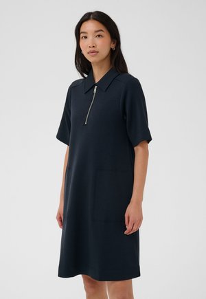 Femme portant une robe bleu marine foncé à manches courtes avec une fermeture éclair devant et une grande poche avant, debout devant un fond uni clair.