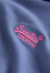 Tela azul marino con una textura suave, que presenta el logotipo bordado en rosa "SuperDry" y un diseño de texto adicional en blanco.