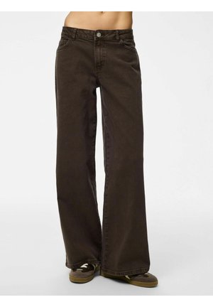 Persoană purtând blugi maro închis cu talie înaltă și croială largă, precum și adidași maro, văzută de la mijlocul trunchiului până la picioare, pe un fundal alb simplu.