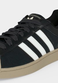 Μαύρο παπούτσι Adidas Superstar με λευκές ρίγες, μπεζ σόλα και μαύρα κορδόνια, εμφανίζεται από μία λοξή πλευρική όψη.