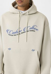 Beige hoodie met trekkoorden heeft blauwe geborduurde tekst "Couture Culture" en golven, met een voorkantzak en ribgebreide accenten.