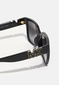 Occhiali da sole neri con montatura spessa, dotati di un logo dorato "MK" sul lato, lenti scure e una finitura lucida.