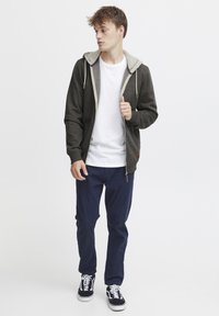 Sudadera con cremallera de color gris oscuro con capucha forrada de felpa beige, combinada con una camiseta blanca básica y pantalones azul marino. Zapatillas de deporte con patrón en blanco y negro.