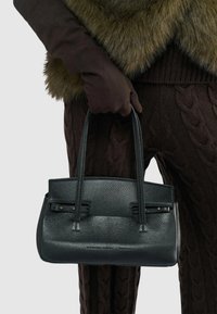 Zwarteleren handtas met een gestructureerde afwerking, twee korte handvatten en subtiele hardwareaccenten. Vastgehouden door een bedekte hand tegen een donkere outfit.