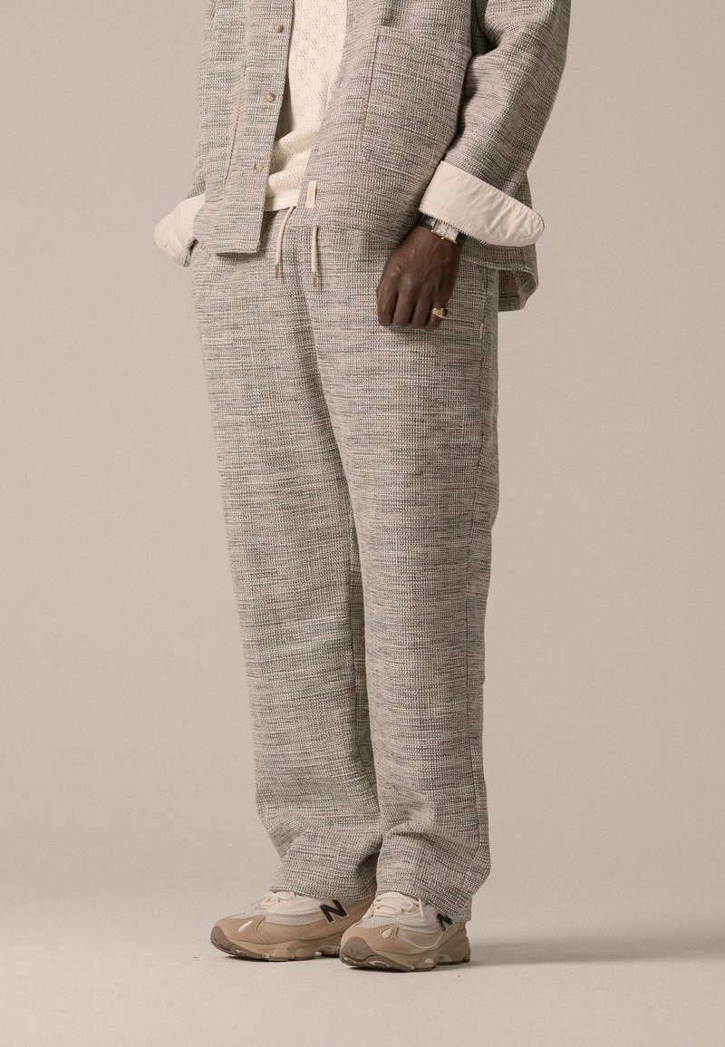 Completo loungewear in tessuto grigio chiaro testurizzato, con giacca con bottoni e pantaloni con cordoncino, abbinato a scarpe da ginnastica beige.