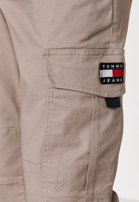 Béžové cargo kalhoty vyrobené z odolné směsové bavlny, s boční kapsou a černou nášivkou s nápisem "TOMMY JEANS" a barevnými pruhy.