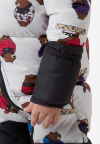 Geacă cu puf pe fundal alb, cu ursuleți de desene animate în diverse culori. Mâneci negre cu manșete elastice. Față cu fermoar.