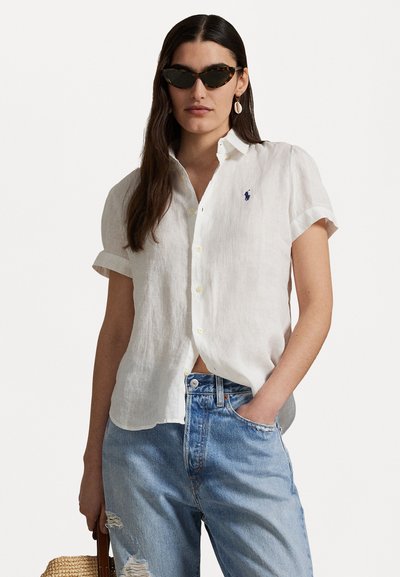 Camisa de lino blanca de botones con mangas cortas remangadas, que presenta un pequeño logo azul, combinada con unos vaqueros desgastados de un azul claro.