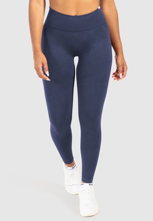 Marineblauwe high-waisted leggings van soepel, rekbaar materiaal met een geribde tailleband en gestructureerde accenten langs de dijen. Geroeid met witte sneakers.