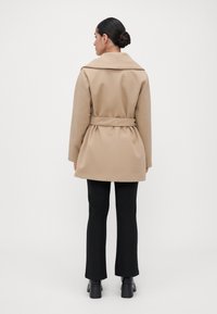 Kvinne med mørkt hår i en knute, iført en beige belted frakk, svarte bukser og svarte hæler, som står med ryggen til mot en enkel bakgrunn.