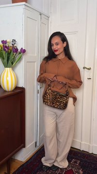 Femme en blouse marron et pantalon beige tenant un sac à motif léopard, debout près d'un placard blanc et d'un vase rayé jaune avec des fleurs violettes.