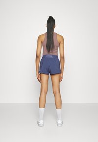 Mujer de pie de espaldas, vistiendo una camiseta sin mangas color malva, pantalones cortos deportivos azul marino, calcetas blancas y zapatillas blancas, frente a un fondo liso.