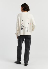 Cremefarbener Pullover mit schwarzer Stickerei, die eine Figur, verstreute Formen und ein Hundedesign zeigt. Kombiniert mit dunkelgrauen, geraden Jeans.
