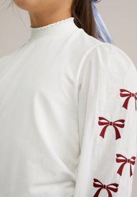 Balta blūze ar augstu smalki noberztu apkakli, pūkainām piedurknēm un sarkaniem izšūtiem loku motīviem uz piedurknēm, izceļot maigu kokvilnas struktūru.