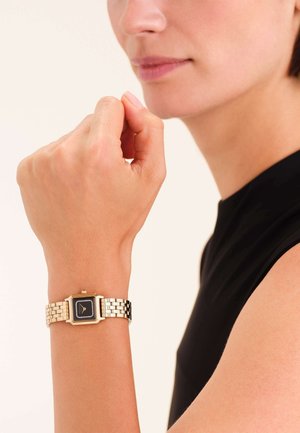 Orologio da polso in tonalità oro e argento con quadrante quadrato nero, cinturino texture e accenti rettangolari, indossato su una mano con uno sfondo neutro.