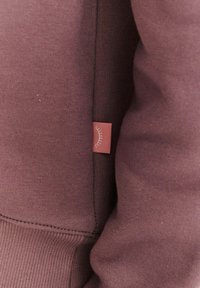 Bordowa bluza z miękkiej tkaniny, z boczną metką ozdobioną prostym wzorem linii. Projekt obejmuje ściągacze na mankietach i u dołu bluzy.