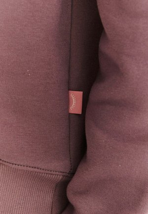 Bordowa bluza z miękkiej tkaniny, z boczną metką ozdobioną prostym wzorem linii. Projekt obejmuje ściągacze na mankietach i u dołu bluzy.