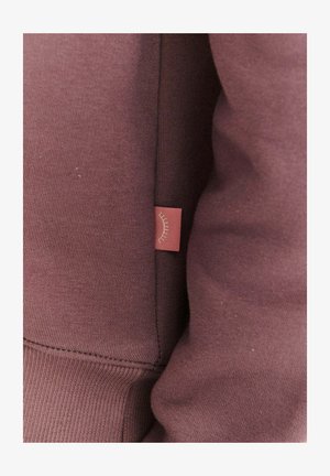 Bordowa bluza z miękkiej tkaniny, z boczną metką ozdobioną prostym wzorem linii. Projekt obejmuje ściągacze na mankietach i u dołu bluzy.