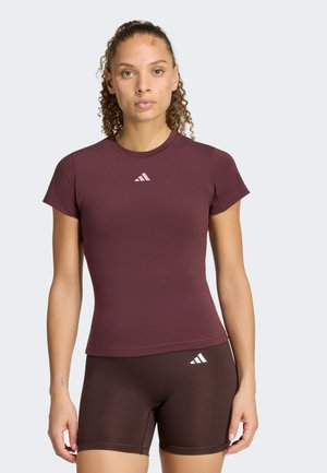 Vrouw met krullend haar draagt een aansluitend donkerrood T-shirt met korte mouwen en bijpassende korte broek met Adidas-logo op de borst en dij, staand tegen een effen achtergrond.