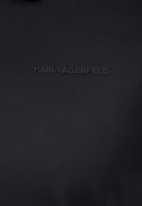 Černá elastická tkanina s hladkou texturou, ve středu zdobená prohloubeným logem "KARL LAGERFELD" pro účely brandingu.
