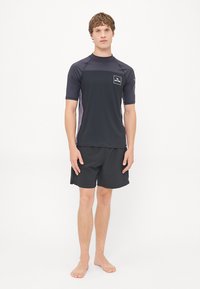 Rövid ujjú fekete rash guard színes panelekkel és logóval, fekete csíkos úszóshortsával párosítva. Az anyag könnyűnek és rugalmasnak tűnik.