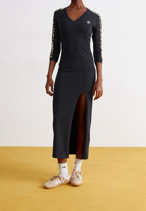 Mujer con vestido negro de manga larga y escote en V con rayas de estampado animal en las mangas, abertura en el muslo, calcetines blancos y zapatillas Adidas beige.