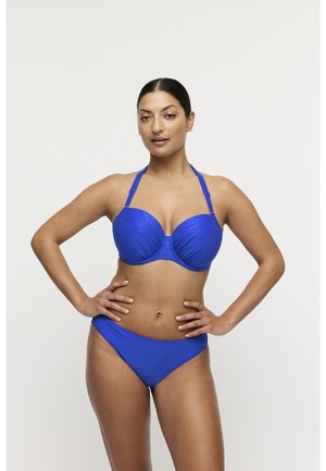 Vrouw staat met handen in de zij, draagt een blauwe gebreide halterbikini tegen een effen lichte achtergrond.