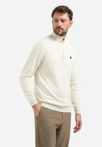 Lichtcrème pullover trui met een halve rits hals, lange mouwen en geribbelde manchetten. Heeft een klein logo op de borst. Gemaakt met beige broeken.