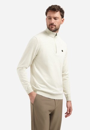 Heller cremfarbener Pullover mit halbem Reißverschluss, langen Ärmeln und gerippten Bündchen. Mit einem kleinen Logo auf der Brust. Getragen mit beigen Hosen.