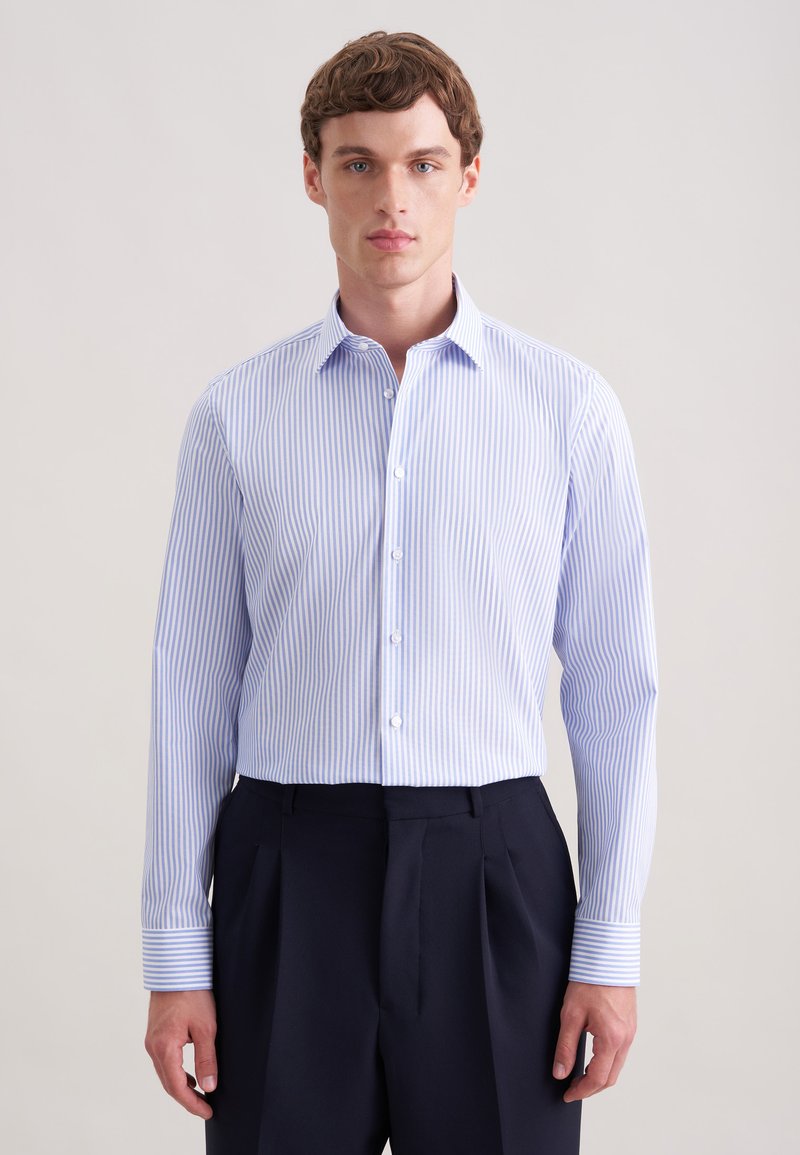 Jeune homme de face portant une chemise à rayures verticales bleu clair et blanc à boutons et un pantalon sombre sur un fond uni.