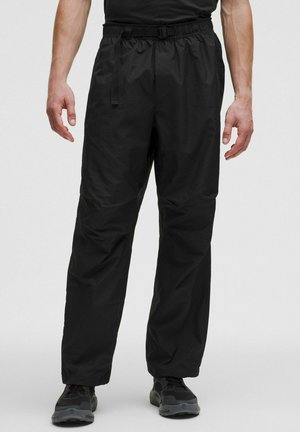 Mand iført løse sorte bukser med justerbar spænde i taljen og sorte sportssko, stående på en lysegrå baggrund.