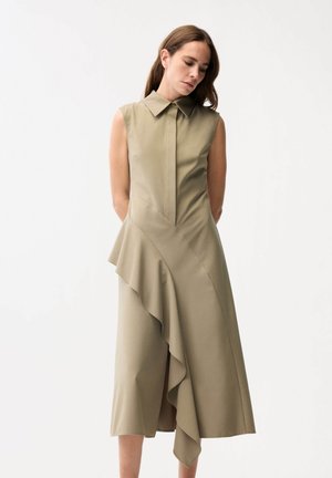 Frau mit langen braunen Haaren, die ein ärmelloses beiges Kleid mit Kragen und asymmetrischem Rüschendetail trägt, steht vor weißem Hintergrund.