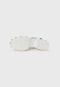 White footwear with a textured sole featuring a rugged design and small black studs along the edges. Clear traction patterns on the sole.