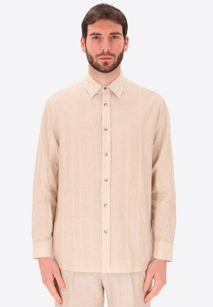 Uomo con barba che indossa una camicia beige a maniche lunghe con bottoni e pantaloni beige abbinati, in piedi su uno sfondo chiaro e uniforme.