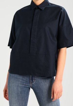 Osoba nosząca luźną, krótka granatową koszulę z kołnierzykiem i plisą z przodu, zestawioną z niebieskimi jeansami.