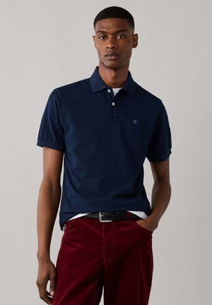 Jonge man die een donkerblauw poloshirt met een klein logo en een rode broek draagt, staat met één hand in zijn zak tegen een effen achtergrond.