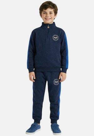 Garçon souriant, portant un survêtement bleu marine avec des rayures bleu clair, un logo sur la poitrine et la cuisse, ainsi que des baskets bleues assorties, debout devant un fond blanc.