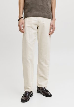 Personne portant un pantalon droit couleur crème, des mocassins foncés polis avec des pompons et un haut en maille marron à manches courtes, debout sur fond blanc.