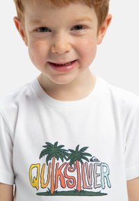 Camiseta de algodón blanca con el colorido logo de "Quiksilver" acompañado de palmeras. Cuello redondo simple, textura suave y elementos de diseño vibrantes.