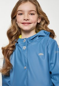 Kék vízálló dzseki kapucnival, ezüst színű patent gombokkal és világos sárga béléssel. A mellkasán egy kis ernyőlogó található.