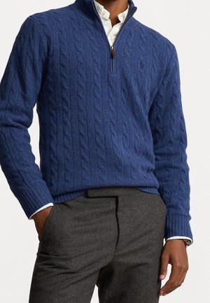 Mann trägt blauen Strickpullover mit Reißverschluss am Kragen über weißem Hemd und grauer Hose, linke Hand in der Tasche und rechte Hand entspannt.