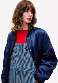 Maritime satijnen bomberjack over een rode geribbelde top, gecombineerd met blauwe gestreepte overalls. Voorzien van een ritssluiting en zijzakken op de overalls.