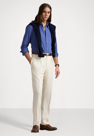 Camisa azul-clara de botão, suéter de malha azul-marinho colocado sobre os ombros, calças creme, sapatos de couro castanho e um cinto castanho.