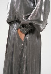 Abito lungo di un grigio metallico con vita regolabile, maniche a palloncino e tasche laterali con dettagli di cucitura sottili. Texture liscia e riflettente.