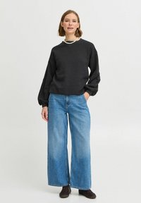 Felpa grigia scura con maniche lunghe, abbinata a jeans a gamba larga blu chiaro. Il modello indossa una semplice collana di perle e scarpe marroni.